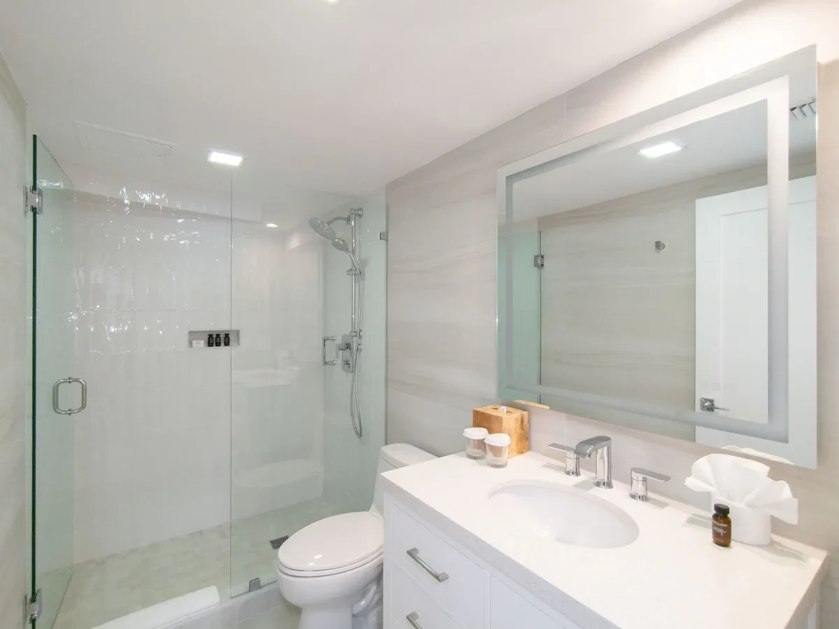 Boutique hotel bathroom at Bentley Hotel South Beach — frameless glass walk-in shower, marble-look tile walls, white vanity with chrome hardware and backlit mirror, IV Atelier Miami Beach hotel design