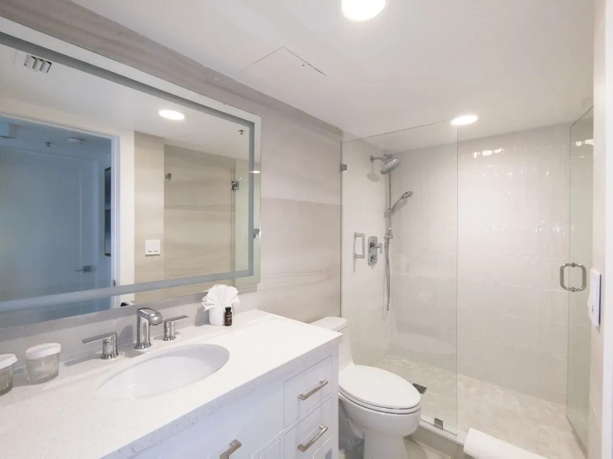 Renovated boutique hotel bathroom at Bentley Hotel South Beach — white quartz vanity, large format neutral tile walls, frameless glass shower with rain head, chrome fixtures, IV Atelier South Beach renovation