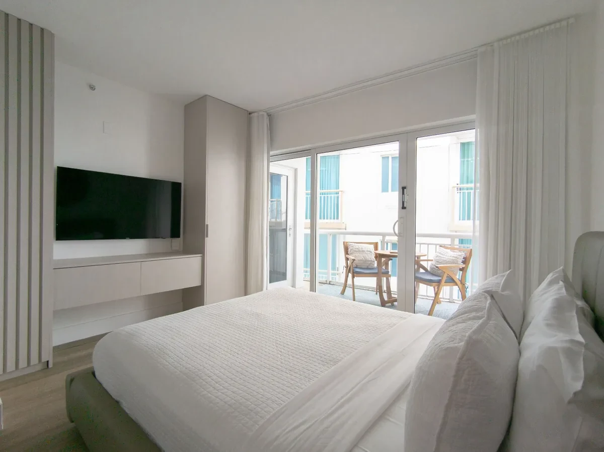 Boutique hotel bedroom at Bentley Hotel South Beach — grey striped panel TV wall, floating white console, white bed with balcony view, IV Atelier South Beach renovation Ocean Drive