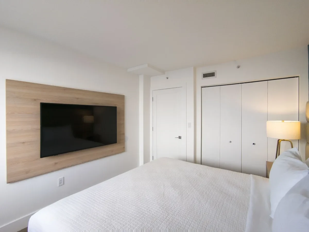 Boutique hotel bedroom at Bentley Hotel South Beach — oak TV wall panel, white bed with crisp white linens, built-in wardrobe, gold tripod lamp, IV Atelier interior design Miami Beach