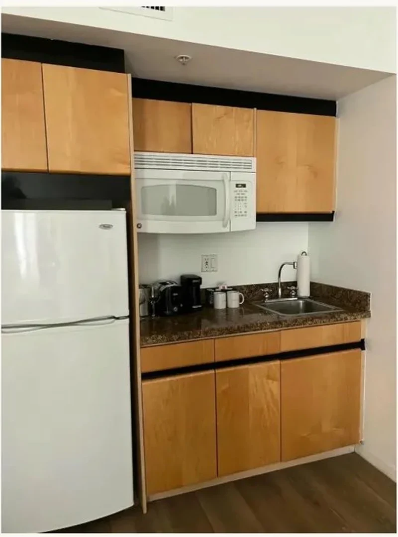 Before renovation — Bentley Hotel South Beach kitchenette with dated oak cabinetry, white refrigerator, white microwave and dark granite countertop, prior to IV Atelier redesign