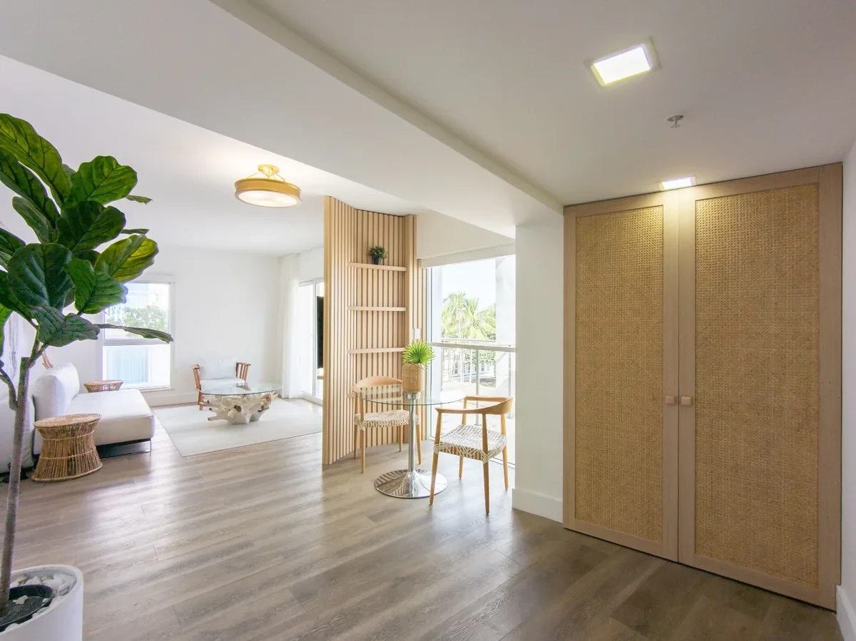 Bentley Hotel South Beach suite entry — rattan cane wardrobe doors, slatted wood room divider, open-plan living with glass dining table and rattan chairs, fiddle-leaf fig tree, IV Atelier Ocean Drive renovation