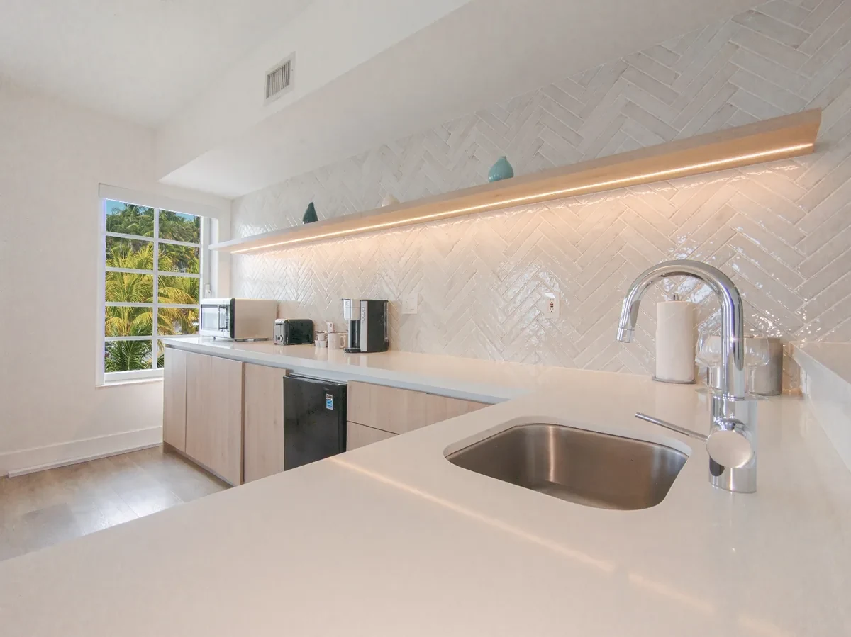 Renovated boutique hotel kitchenette at Bentley Hotel South Beach — white gloss herringbone tile backsplash with LED shelf lighting, light oak flat-panel cabinetry, quartz countertop and palm garden view, IV Atelier
