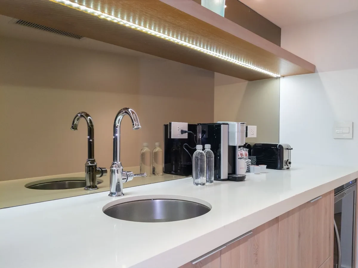 Close detail of boutique hotel kitchenette at Bentley Hotel South Beach — dual round undermount sinks with gooseneck chrome faucets, white quartz countertop, LED-lit oak shelf above, IV Atelier South Beach renovation