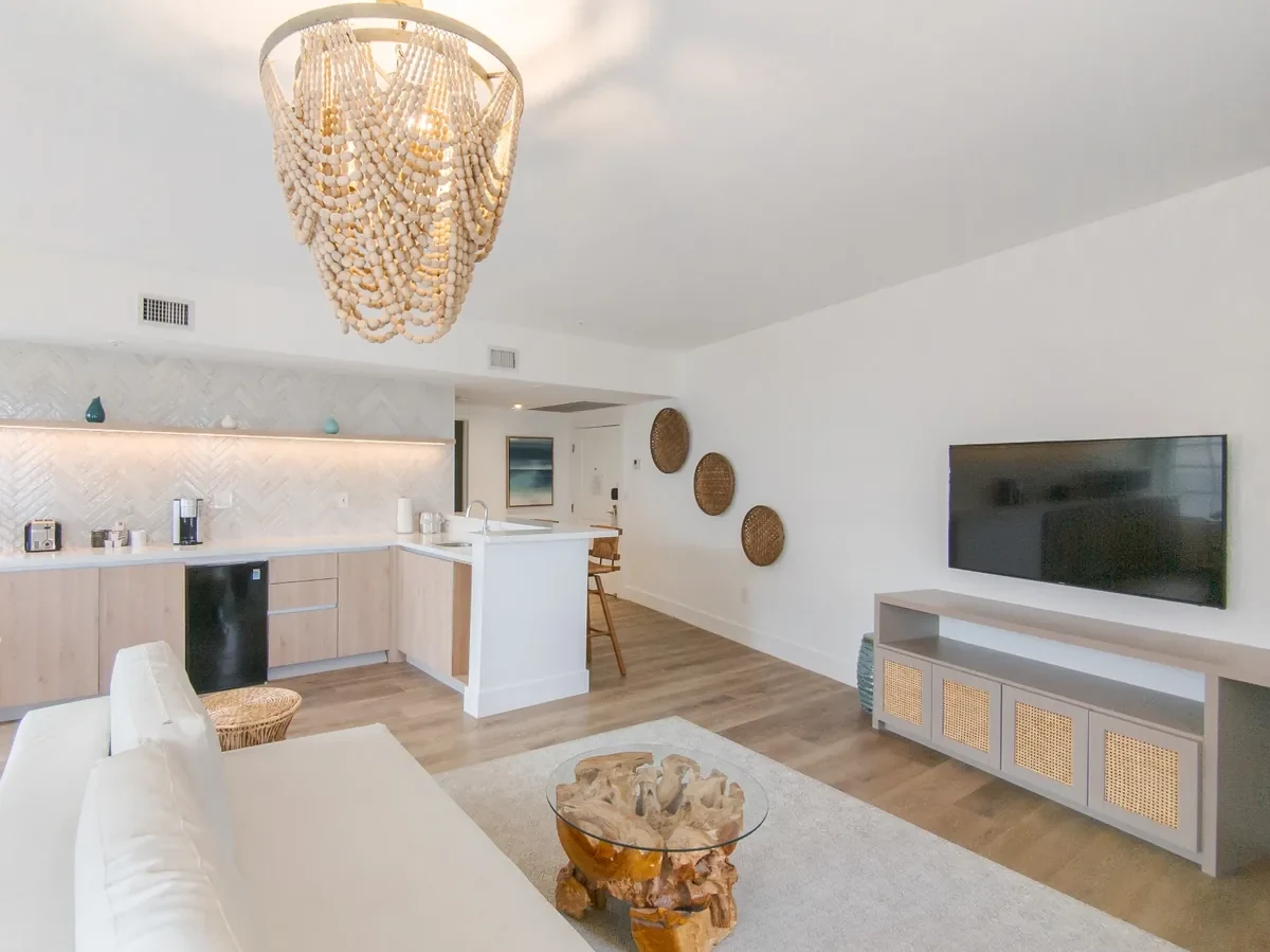 Open-plan boutique hotel suite at Bentley Hotel South Beach — beaded shell chandelier, herringbone tile backsplash kitchenette, rattan media console, woven wall art and teak root coffee table, IV Atelier interior design