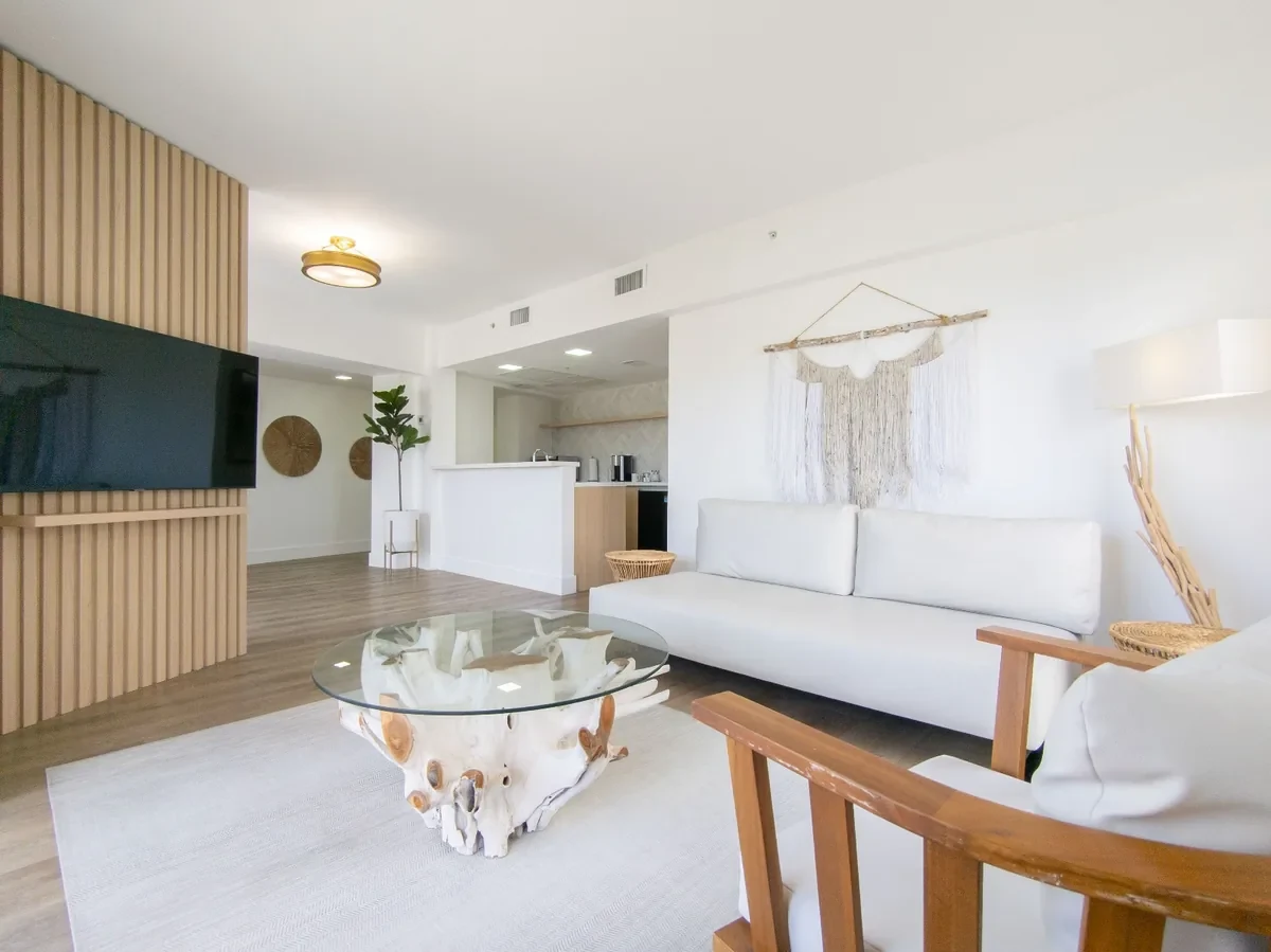 Boutique hotel suite at Bentley Hotel South Beach — slatted wood TV wall, macramé wall hanging, teak root coffee table, kitchen peninsula beyond, gold flush mount ceiling light, IV Atelier Miami Beach renovation