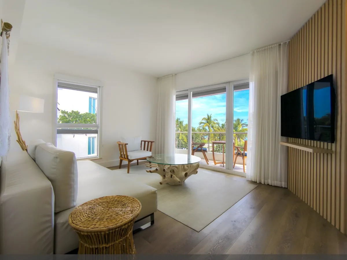 Boutique hotel suite living room at Bentley Hotel South Beach — white sofa, teak root glass coffee table, slatted wood TV wall and floor-to-ceiling sliding doors opening to ocean-view balcony, IV Atelier South Beach