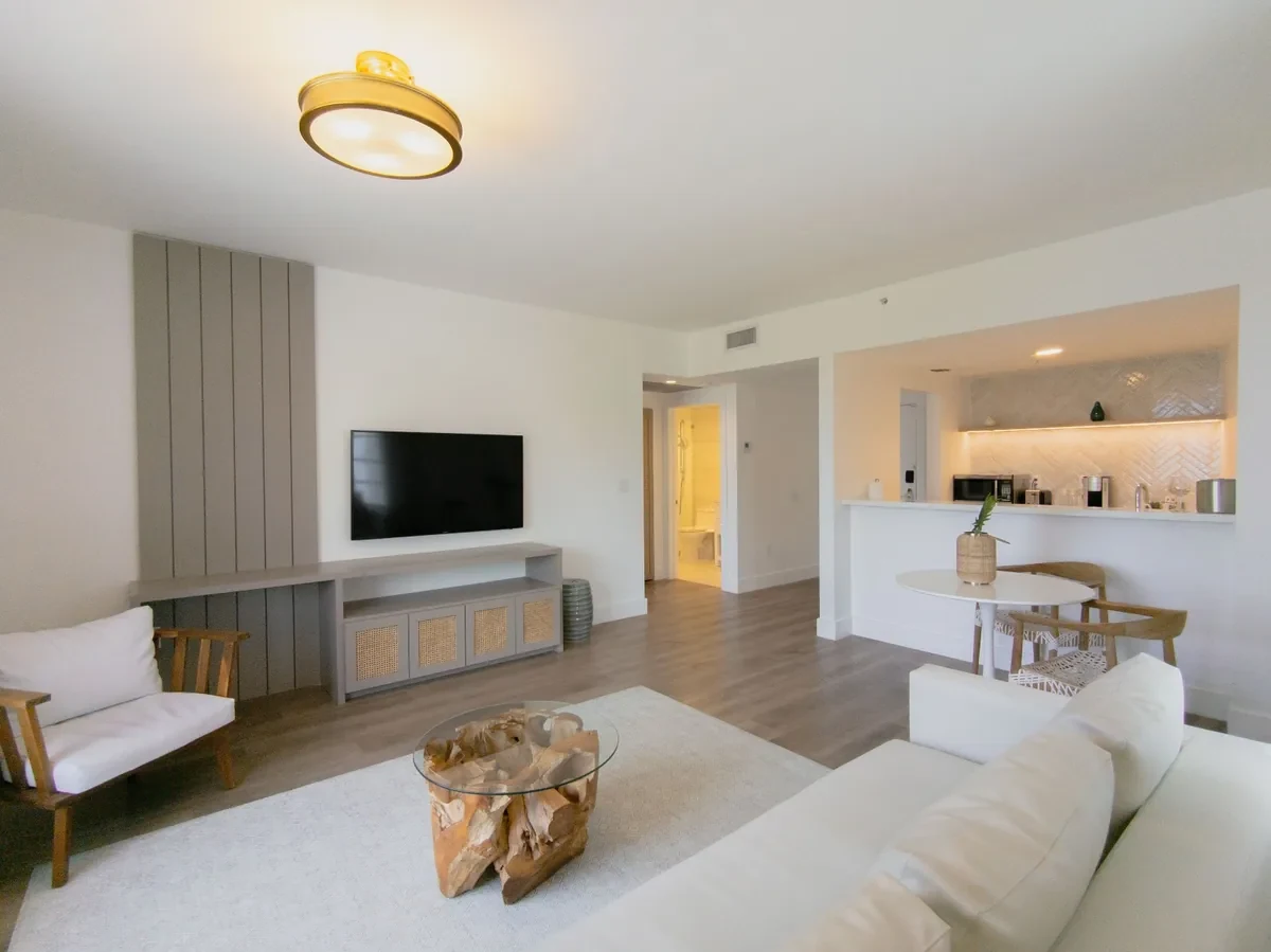 Boutique hotel open-plan suite at Bentley Hotel South Beach — grey panel TV wall, rattan cane console, teak root coffee table, kitchen peninsula with herringbone backsplash visible beyond, IV Atelier Miami