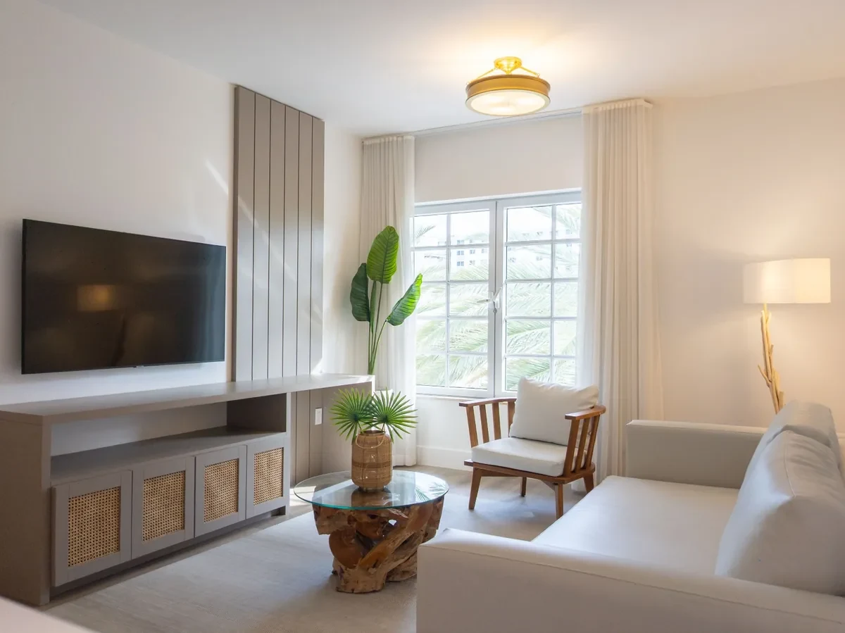 Boutique hotel suite at Bentley Hotel South Beach — grey panel TV wall, rattan cane console unit, teak root coffee table, bird of paradise plant and large window with palm view, IV Atelier South Beach