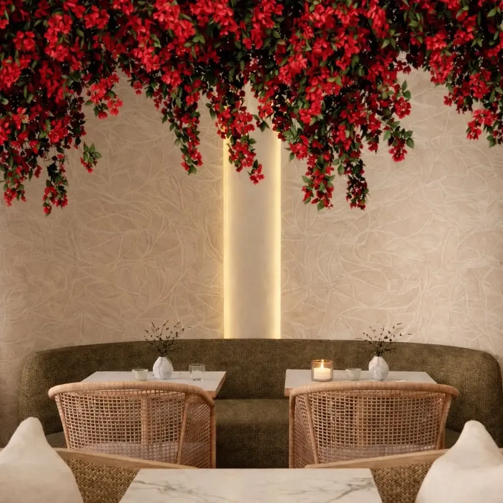Intimate restaurant dining room design South Beach Miami — lush cascading red bougainvillea floral canopy above curved olive bouclé banquette, handwoven cane dining chairs, white ceramic bud vases and candlelit marble-top tables beneath warm textured plaster wall with illuminated LED reveal, IV Atelier Brisa Casa Ocean Drive